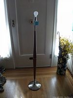 Full view of the tall teak floor lamp standing near a door, showing its height, teak wooden shaft, and metal base, with light bulb installed but no shade.