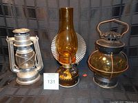 Three oil lamps: new galvanized metal storm lantern on left, amber glass hurricane lamp with metal reflector in middle, round metal and glass lantern with handle on right.