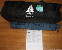 Black Woods Jasper 6 tent bag, blue storage bag, and owner manual on wooden floor.