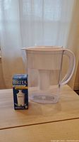 Photo showing Brita water filter pitcher with white lid and handle beside a box containing one replacement Brita water filter.
