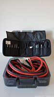 Booster cables coiled inside a gray plastic carrying case with a black roll-up tool pouch laying open behind it showing the small tools.