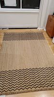 Full view of entire jute rug laid flat on hardwood floor, showing natural color and dark chevron zigzag painted pattern on both edges.