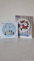 Two cross stitch kits side by side, one with a floral bouquet design and the other titled Summer Weekend Floral Bouquet.