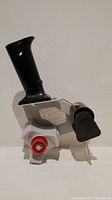 Front and angled side view of the tape dispenser showing black plastic handle, metal frame, and cutter blade with tension adjustment knob.