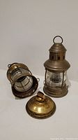 Pair of brass and glass lanterns, one laying on side with bottom twisted off revealing inner reservoir and wick mechanism.
