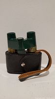 Green small binoculars with brass details standing on their leather carrying case with a wrist strap