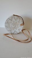 Full front view of the round purse showing floral white woven pattern and leather strap laid out on surface.