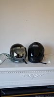 Two motorcycle helmets side by side on a mantel, one black Shoei and one silver Bell helmet with visors up.