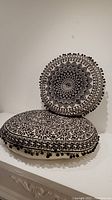 Two round Indian cushions, one standing and one lying flat, showing full view of black floral design on cream fabric with pom-pom trim.