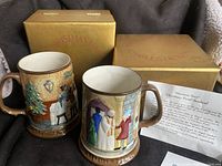 Two ceramic Beswick Christmas Carol mugs with winter scenes, both seated in their original gold boxes with red text indicating the collection name