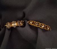 Two gold-tone rings on dark fabric. One shows five clear rhinestones in a row. The other has clear rhinestones encircling it in a decorative pattern.