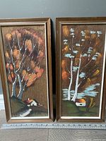 Both paintings framed, showing stylized trees and houses on burlap canvas with visible texture, dated vintage condition