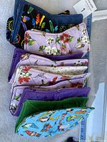 Photo showing 7 folded cotton bowl cozies stacked, revealing different colorful prints including gnomes, hummingbirds, flowers, and dream catchers, alongside a measuring tape showing size about 9 inches square.