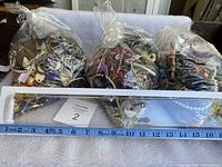 Three bags full of assorted colorful and metallic jewelry pieces including beads, chains, and pendants, placed side by side on a white surface with a measuring tape showing length of about 15 inches.
