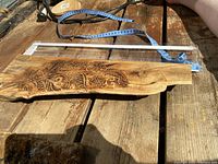 The wood board laying on a wooden table with a tape measure and power cord nearby, showing the length and engraved feather design on the surface.