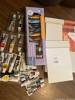 Photo showing acrylic paint tubes in a box, multiple additional acrylic paint tubes outside the box, and canvases stacked together