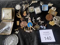 Wide view showing various vintage men's accessories including tie clips, lapel pins, cufflinks, and tie tac pins laid out on a black surface. Items include golf themed tie clip, various lapel pins with medals, a pocket watch or small clock in box, and assorted metal tie clips and pins.