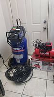 Two air compressors shown together: blue Campbell Hausfeld vertical 22 gallon tank with hoses on floor and red Hyper Tough 3 gallon compressor on box with power cord coiled on top.