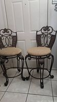 Two ornate metal frame barstools with beige corduroy seats positioned in front of a white door on tiled floor.