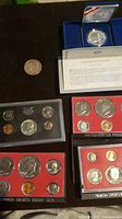 Photo showing two silver dollars including a 1921 Morgan and a 1987 Constitution Silver Dollar with certificate, plus four U.S. proof sets in cases from 1970, 1976, and 1980.