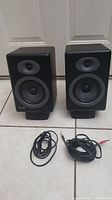 Pair of Audioengine A5 Plus bookshelf speakers side by side with two audio cables placed in front on tile floor, showing overall condition and included accessories.