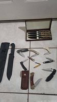 Overview of all knives and sheath showing various folding and fixed blade knives, leather sheath, and 4-piece steak knife set in wood case.