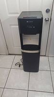 Full frontal view of Glacier Bay water dispenser showing the entire unit with power cord on tile floor in front of closed door.