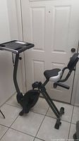 Side view of the black and gray Stamina Wirk recumbent exercise bike with cushioned seat and backrest, positioned in front of a white door on tiled floor.