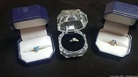 Three gold rings in display boxes: blue heart-shaped gemstone ring, diamond-accented ring in a clear box, and cluster stone ring.