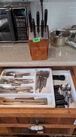 Photo of a wooden knife block containing six black-handled knives and an open drawer with assorted cutlery including forks, spoons, and knives.