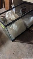 Photo of a rectangular glass fish tank with black trim, containing white packing material and assorted items inside. Set on a concrete floor.
