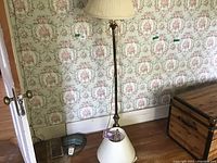 Metal floor lamp with cream-colored lampshade on wooden floor near floral wallpapered wall.