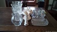 View showing assortment of glassware items on wooden table including two vases, glass decanter, pitcher, small lidded bowl and bowls stacked.