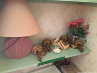 Photo showing pink round Art Deco style lamp base with beige shade, carved wooden shoe, two matching gold curved wall sconces, faux floral decor in metal container, small lidded jar.