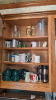 Wooden cabinet shelves holding various mugs, pitchers, beer stein, shot glasses, and a kitchen timer visible at bottom shelf