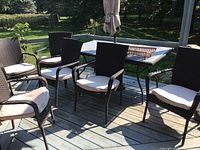 Full view of six brown rattan patio chairs arranged around a glass and metal outdoor table on a wood deck in sunlight.