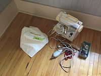 Kenmore sewing machine with cover, foot pedal, cords, fabric scissors, and metal container with accessories placed on wooden floor near wall.