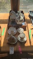 View of assorted porcelain tea cups, saucers, small teapots, and sugar bowl arranged on wooden tray.