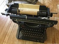 View of the entire vintage black typewriter showing the keyboard, paper roller with a plastic cover rolled inside, resting on a wooden floor and inside a padded noise dampening case.