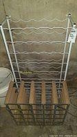 Tall white metal wine rack with horizontal wave-shaped bottle holders for wine bottles, placed against wall.