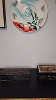 Two Japanese boxes placed side by side on a black surface under a painted wall plate.