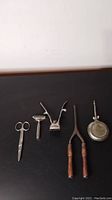 Top view of five antique personal care items arranged horizontally on a black surface showing scissors, shaving device, hair clipper, curling iron, and oil can.