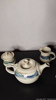 Three-piece ceramic pottery set including teapot, creamer, and sugar container on black surface with plain background.