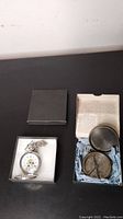 Two boxed items on black surface: silver pocket watch with visible gears in a box and Victorian Trail replica compass with information paper in box.