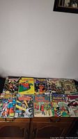 Photo showing 12 various comic books laid out on a wood cabinet, featuring titles such as The Human Fly, Archie, Betty and Veronica, Captain America