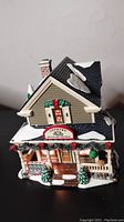 Front view of Department 56 Country Quilts and Pies ceramic house with wreath, snow accents, and small Christmas trees on porch.