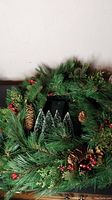 Close-up photo of the circular green Christmas wreath decorated with pinecones, red berries, and two small unattached Christmas trees in the center, unlit LED lights are integrated.
