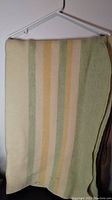 Photo showing the folded antique wool blanket hanging on a white plastic hanger, displaying vertical folds and cream/green/yellow stripes.