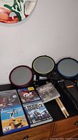 Photo of Rock Band drum set with three colored drum pads and two drumsticks, along with five video game cases laid out.