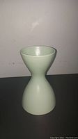 Full side profile of the green hourglass-shaped vase on black surface against plain wall, showing the entire silhouette and surface condition.